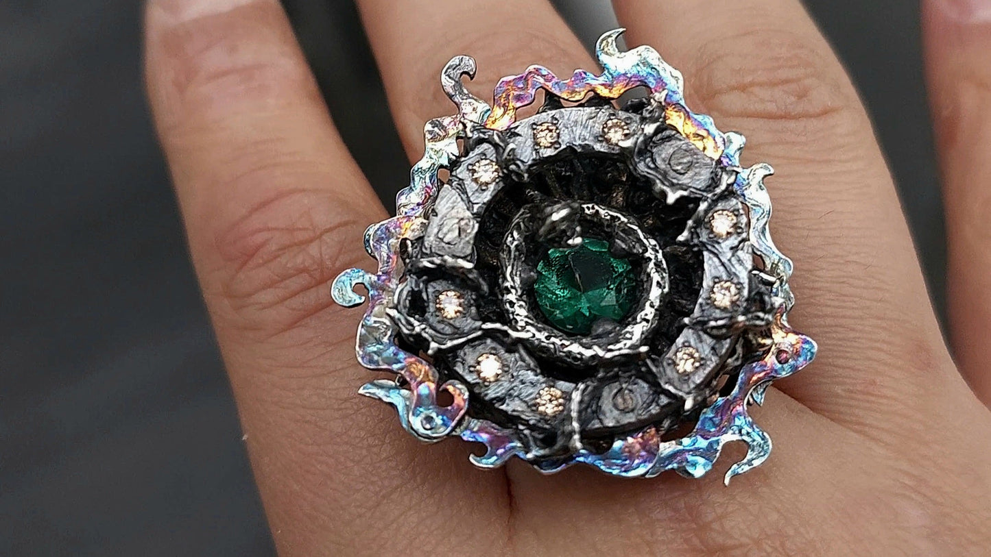 A oxidized ring with iridescent 'poison fog' circling a cavern like design, with snake surrounding and guarding a green quartz within