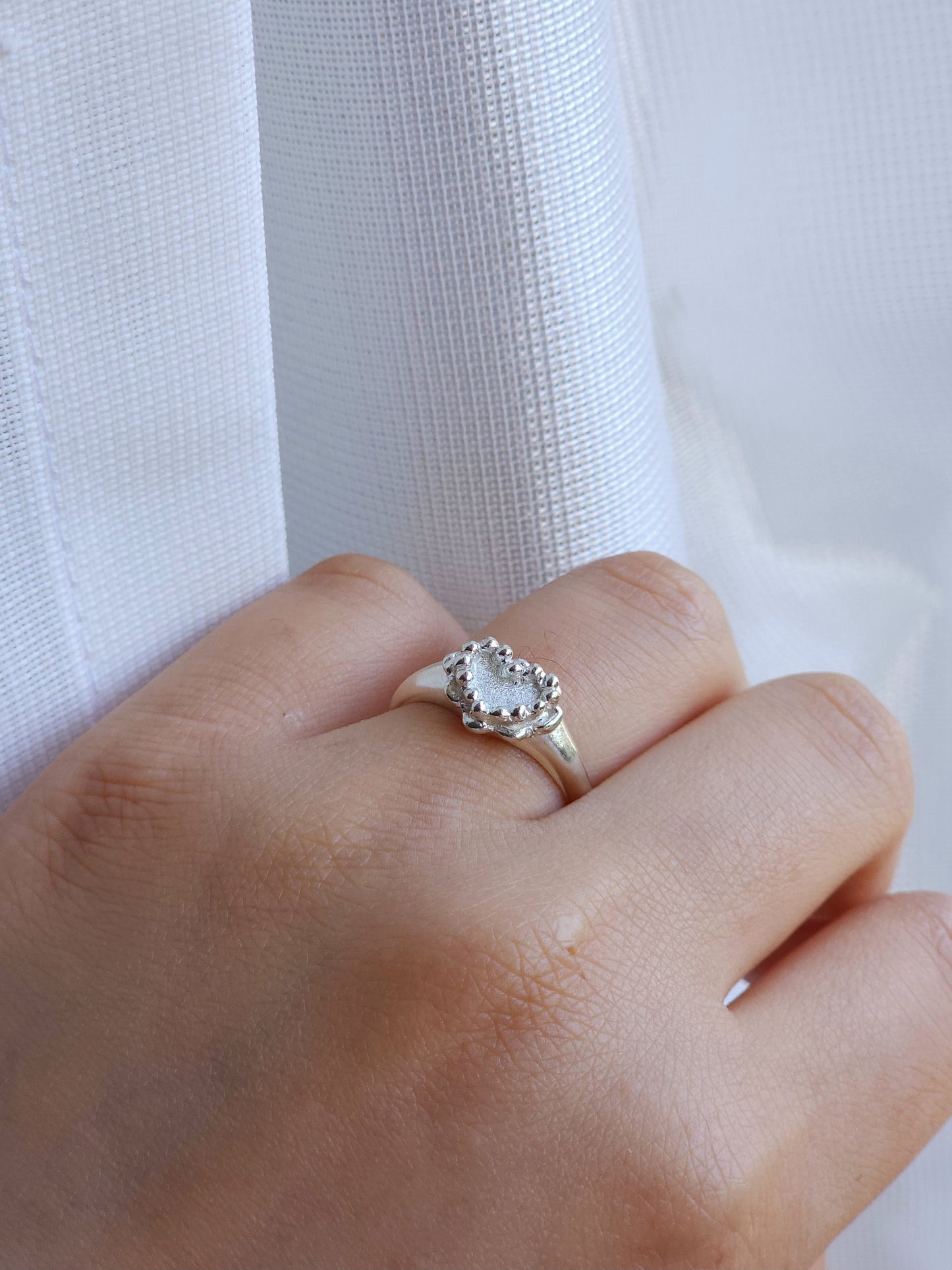 A silver ring with a heart-shaped clear gemstone and a halo of small clear stones, worn on a person's hand against a white textured fabric background.
