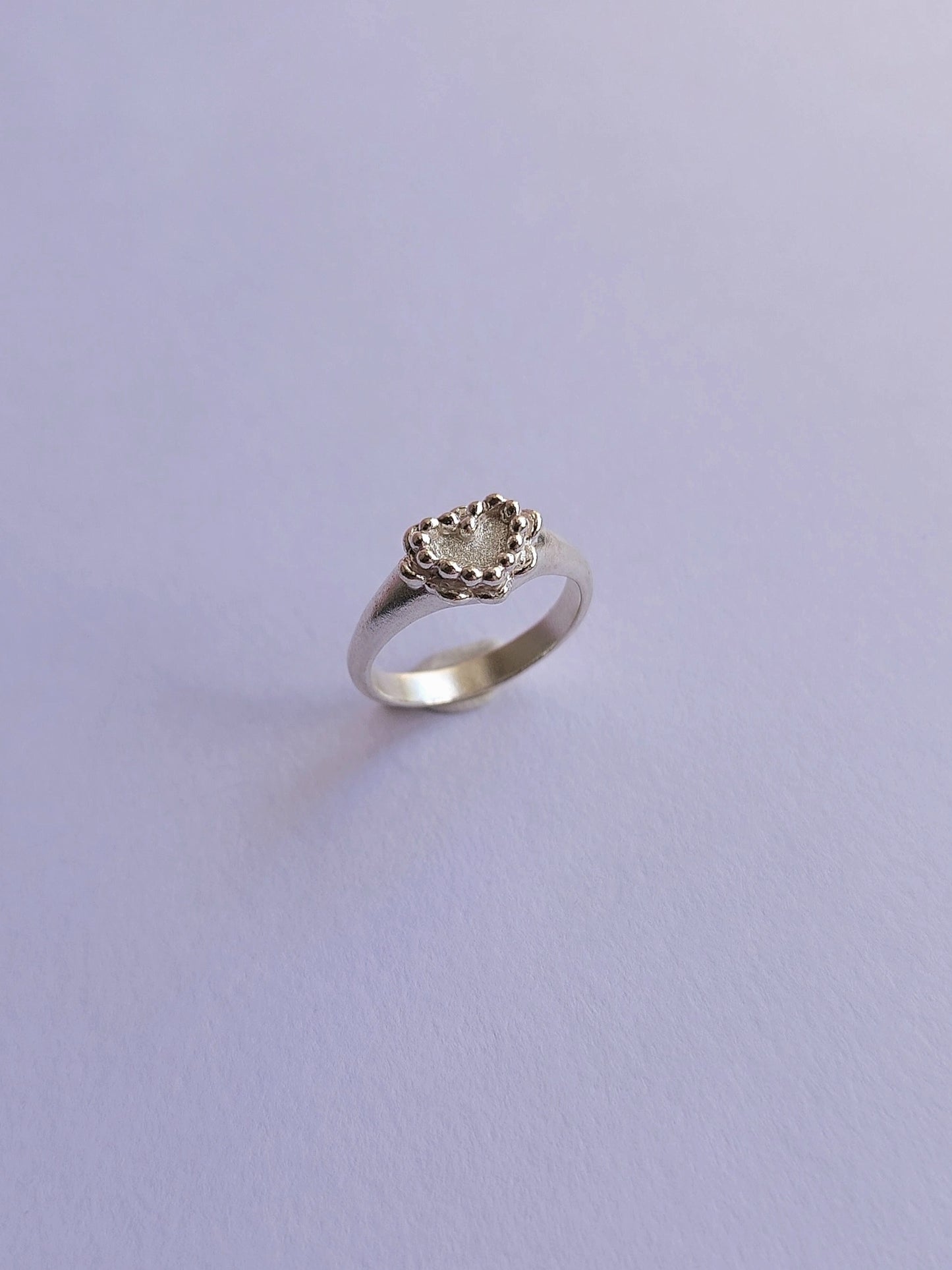 A silver ring with a small heart-shaped floral or leaf cluster setting, photographed on a plain light lavender background.