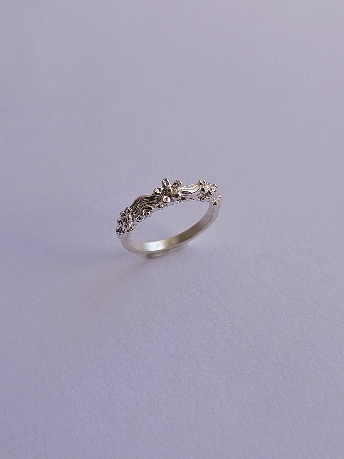 A silver ring with a detailed, textured floral or leafy design on the band, photographed on a plain light gray background.