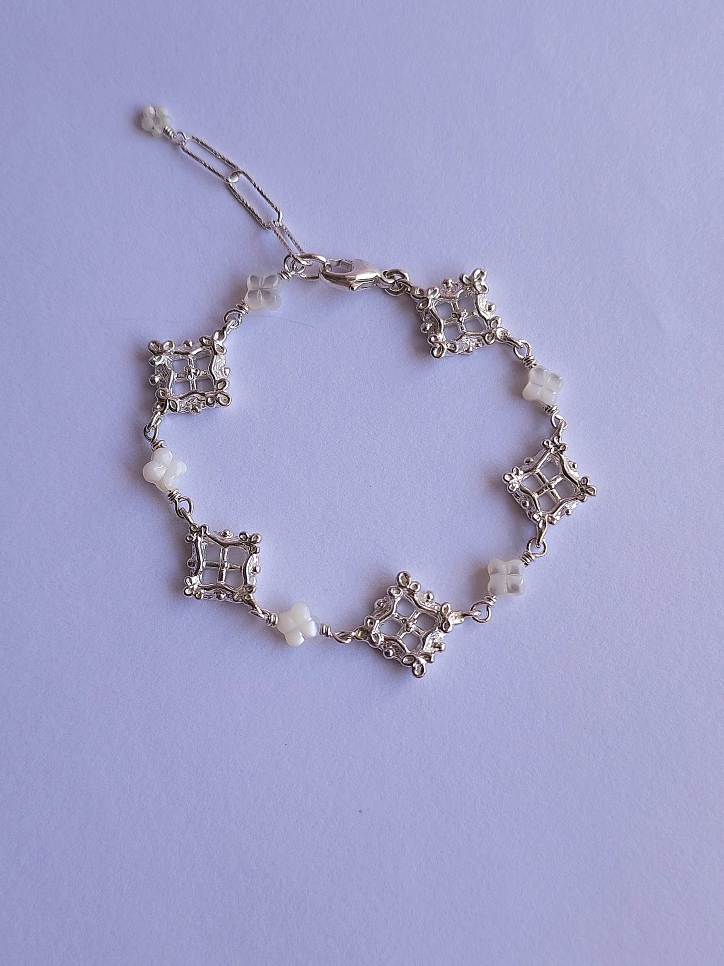 A delicate silver chain bracelet with repeating diamond shaped motifs in an openwork lattice and small white bead accents, laid flat on a light lavender background.