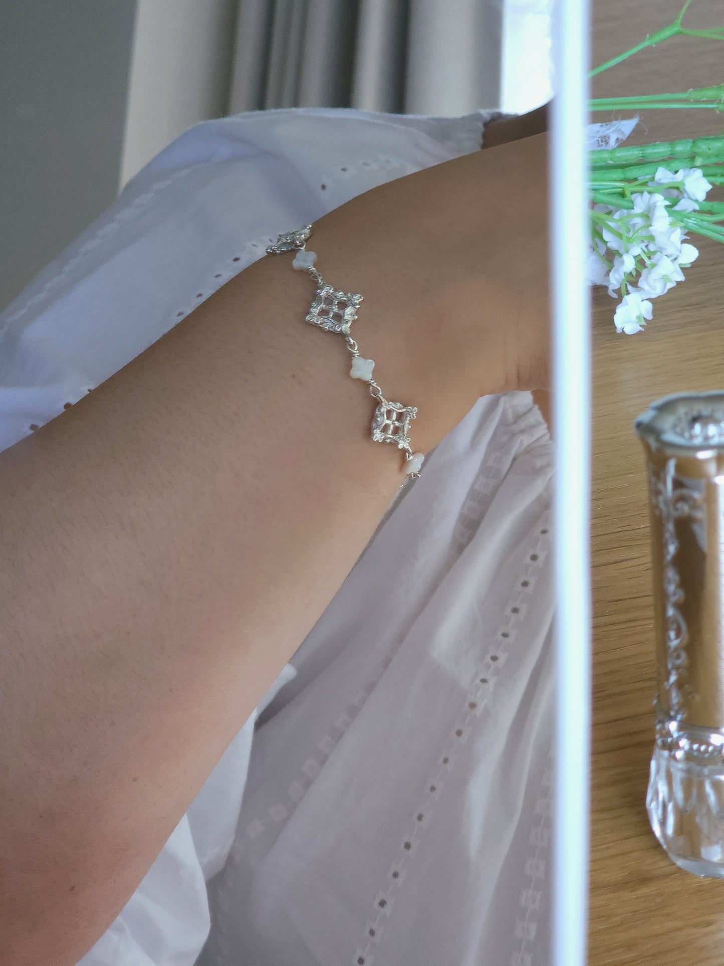 A delicate silver-toned chain bracelet with lattice diamond-shaped links is shown worn on a person's wrist, paired with a white eyelet top. The photo is taken in a softly lit indoor setting reflected in a mirror with a wooden table, flowers, and a ornate lipstick visible in the background.