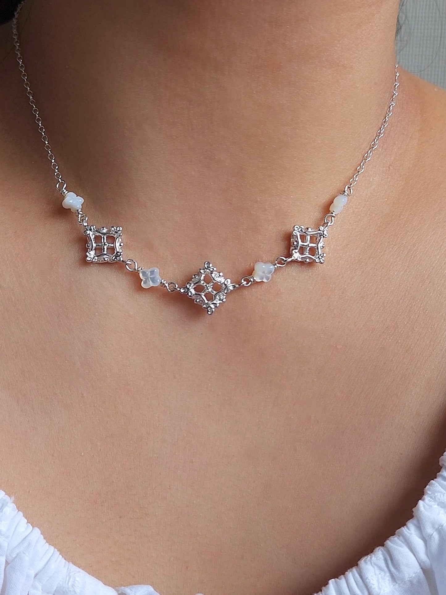 A close-up of a delicate silver necklace with geometric and floral motifs, worn on a model's neck above a white eyelet top.
