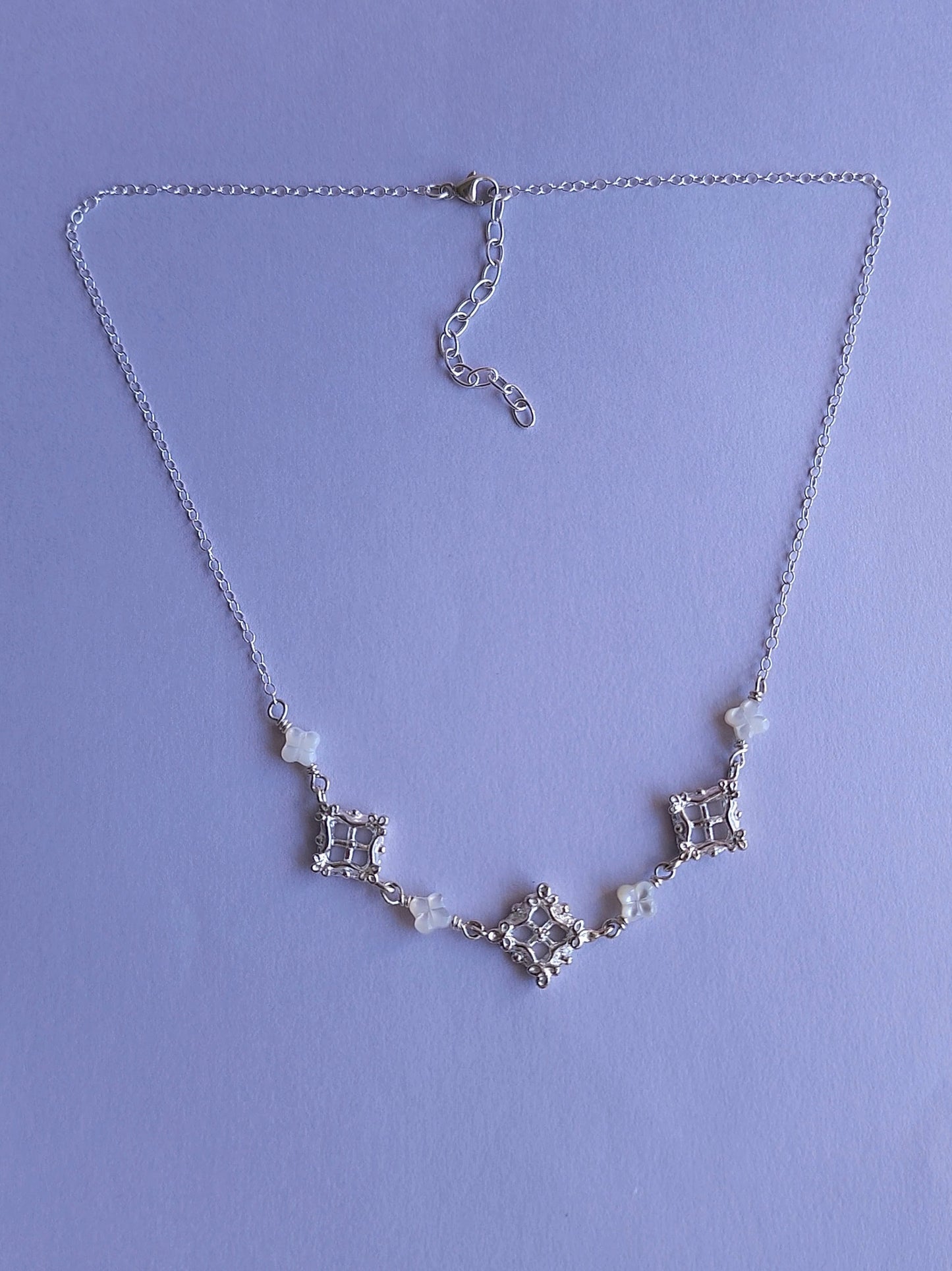 A delicate silver necklace with geometric motifs with a lattice design accentuated with floral details, alternating with white clover-shaped beads, laid on a light purple background.