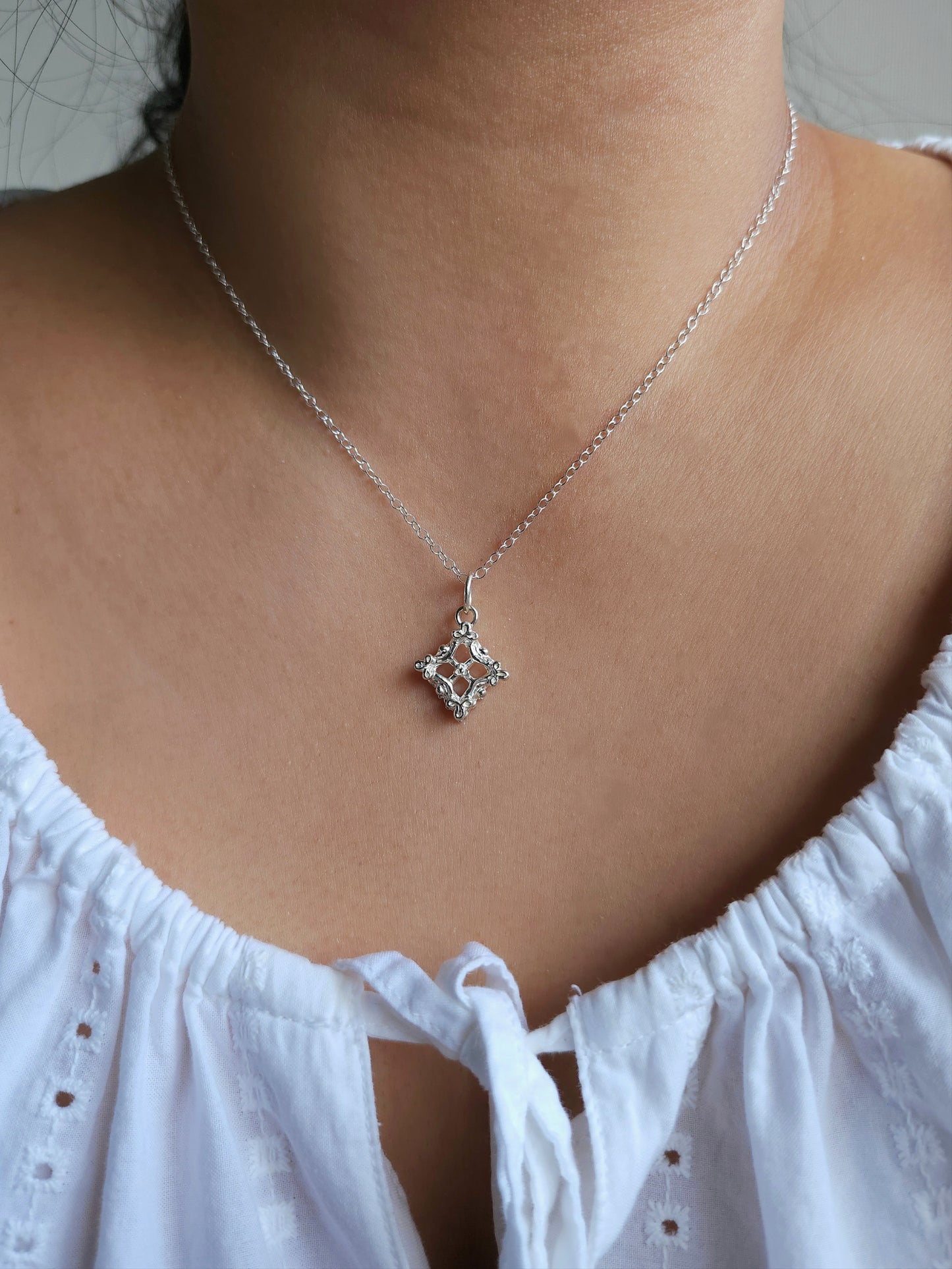A close-up of a person wearing a delicate silver chain necklace with a small geometric diamond-shaped pendant set with clear stones, paired with a white eyelet top.