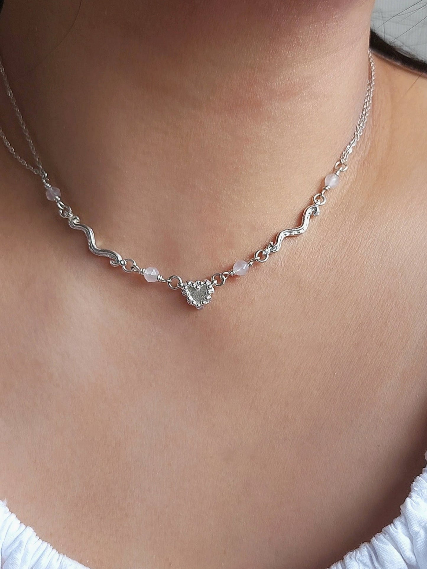 A close-up of a delicate silver wavy chain necklace with a small heart-shaped charm and light pink beads, worn on a model's neck.