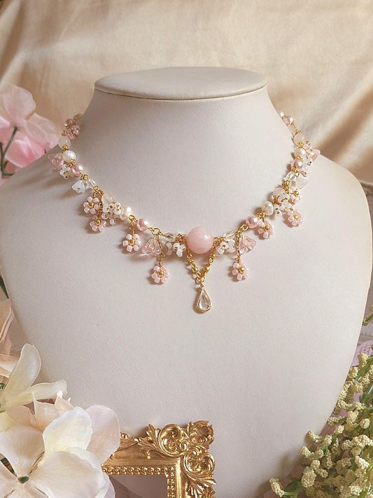 This elegant handmade necklace features rose quartz beads, freshwater pearls, pink glass pearls, and delicate clusters of floral beaded designs in pink and white. Perfect for princess-inspired looks, this romantic jewelry piece showcases a large rose quartz bead centerpiece with a dangling clear droplet for an enchanting touch.