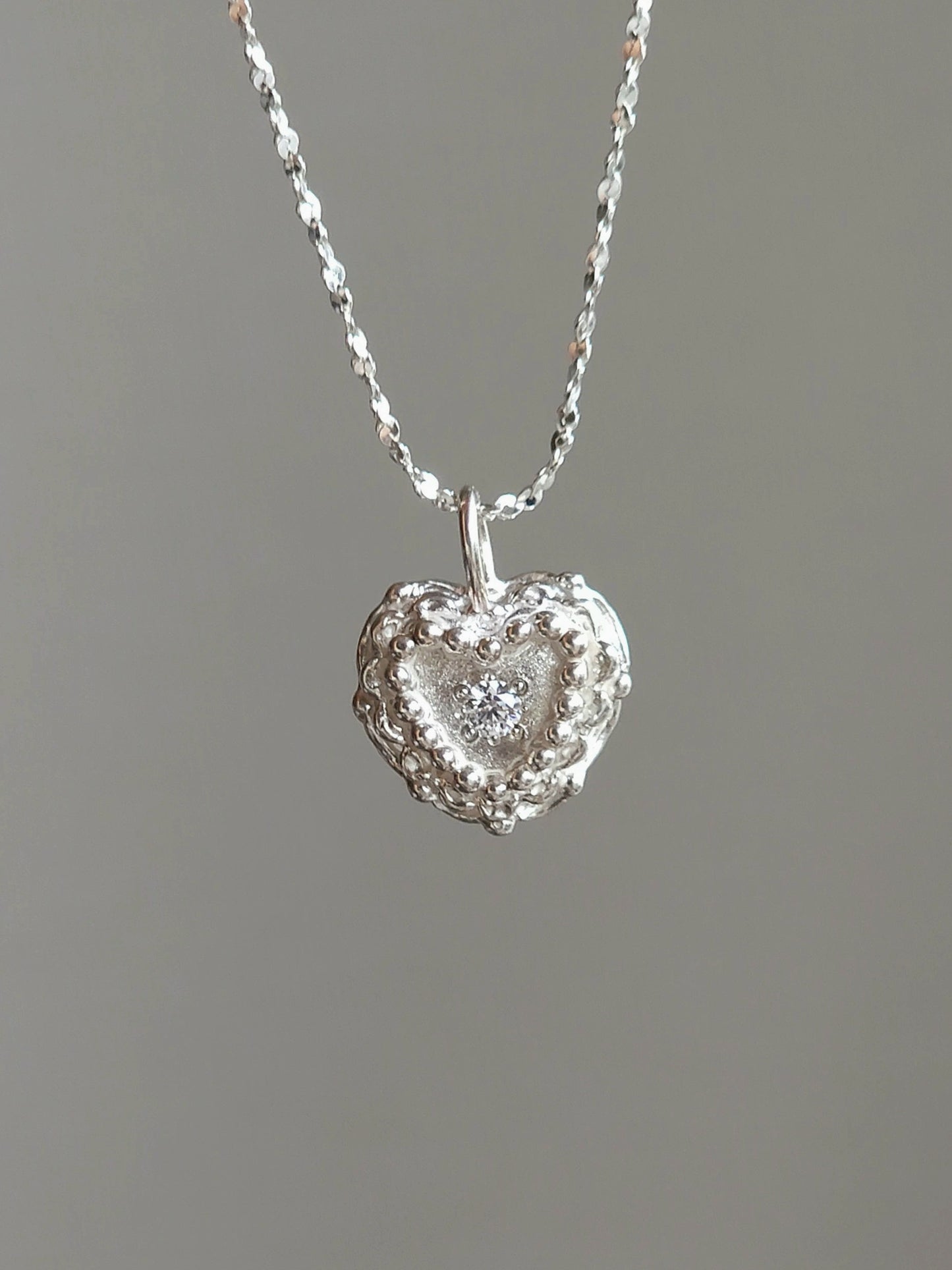 A silver heart-shaped pendant necklace with a textured outer border and a clear central gemstone, suspended from a delicate silver chain against a solid gray background.
