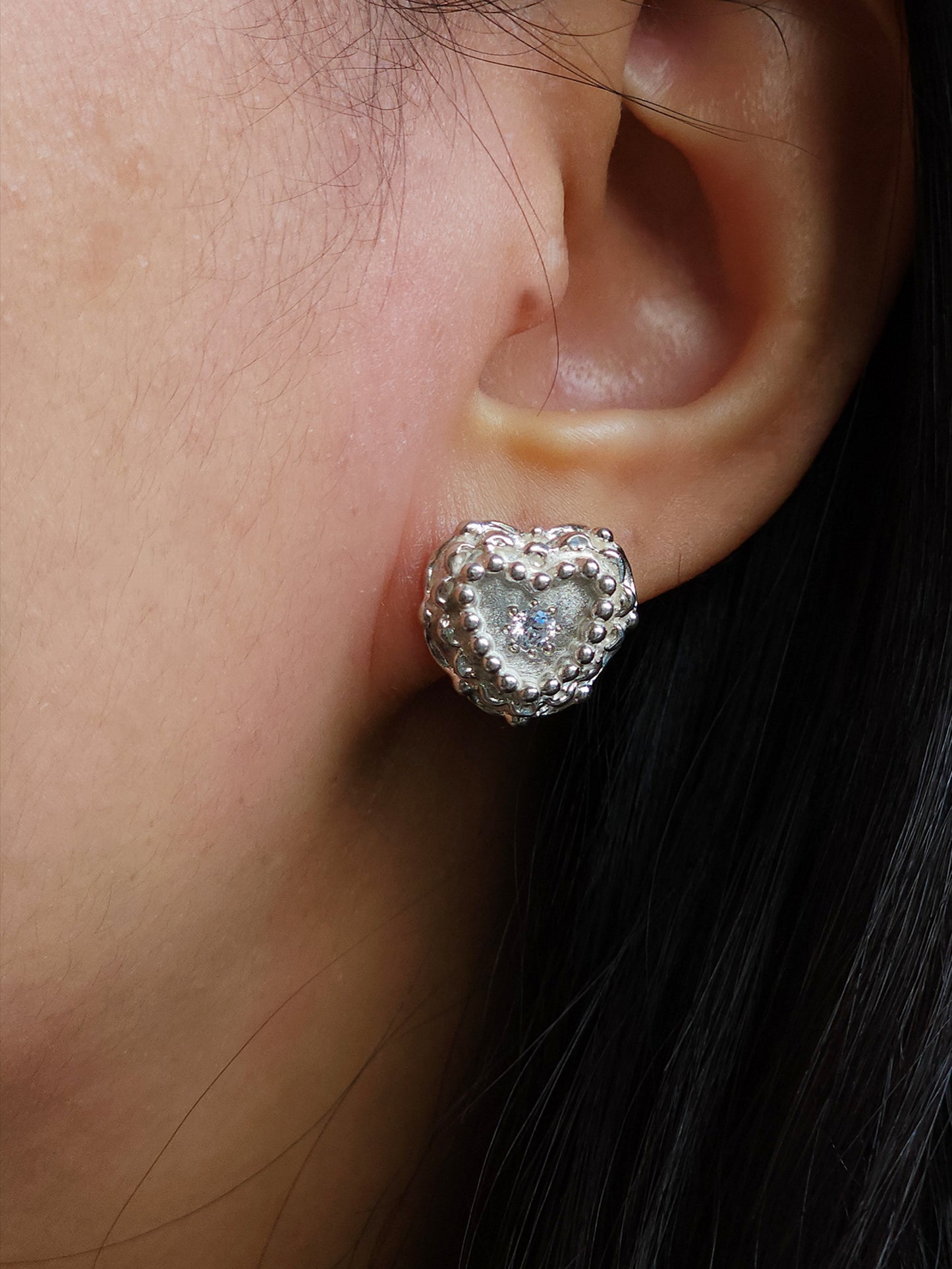 A close-up of a person's ear wearing a heart-shaped silver stud earring with a clear central stone and textured silver border.
