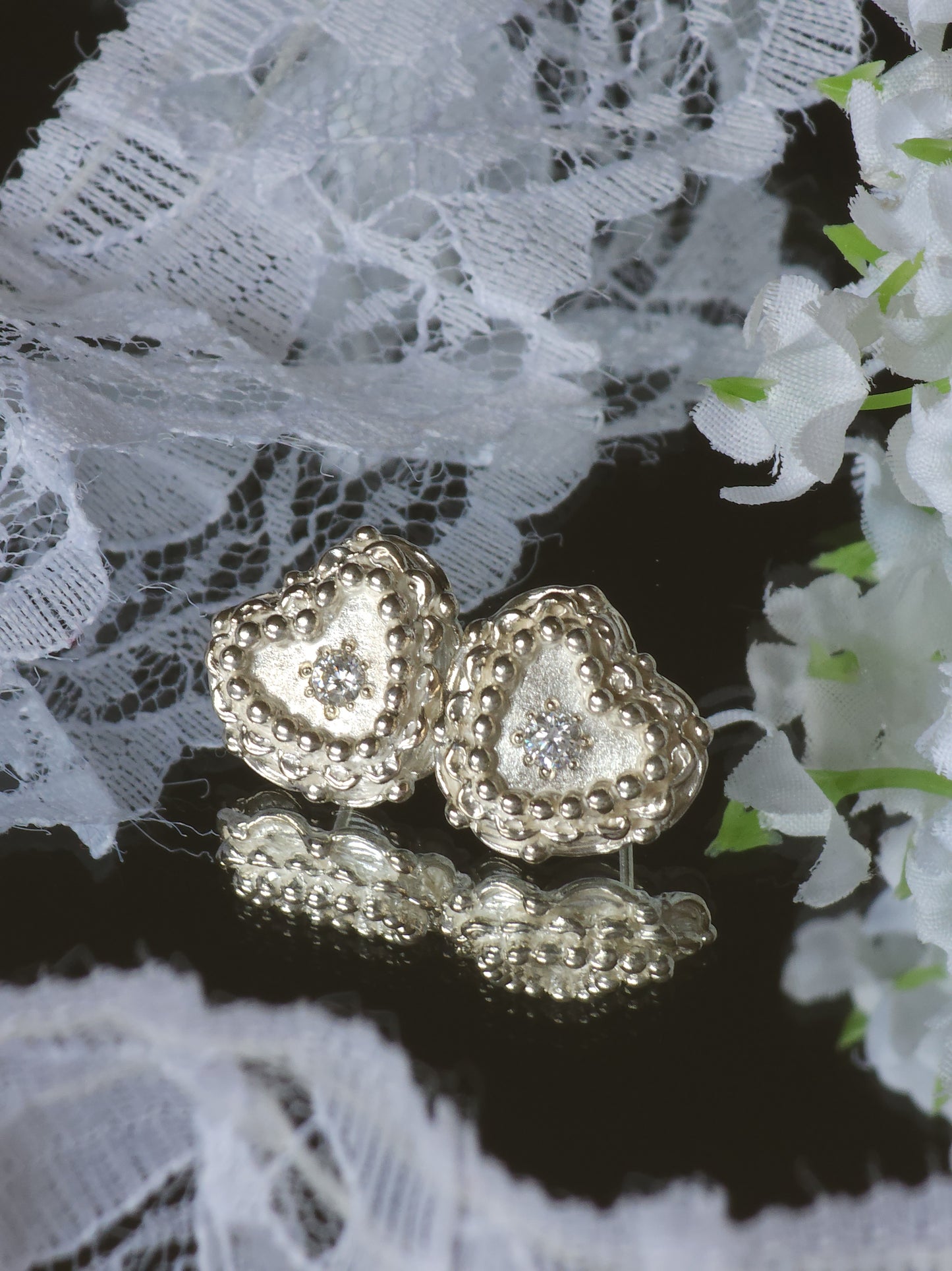 A pair of textured silver heart-shaped earrings with clear gemstones set in the center, displayed on a reflective dark surface with white lace and flowers in the background.