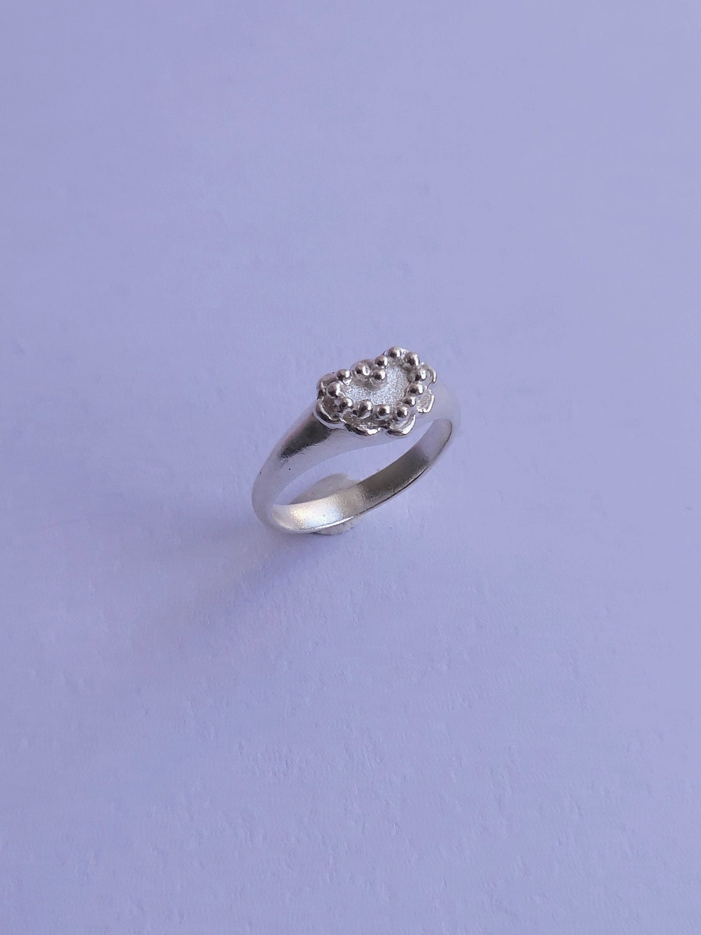 A silver heart shaped signet ring with granulation detail around the perimeter, set on a plain lavender background.