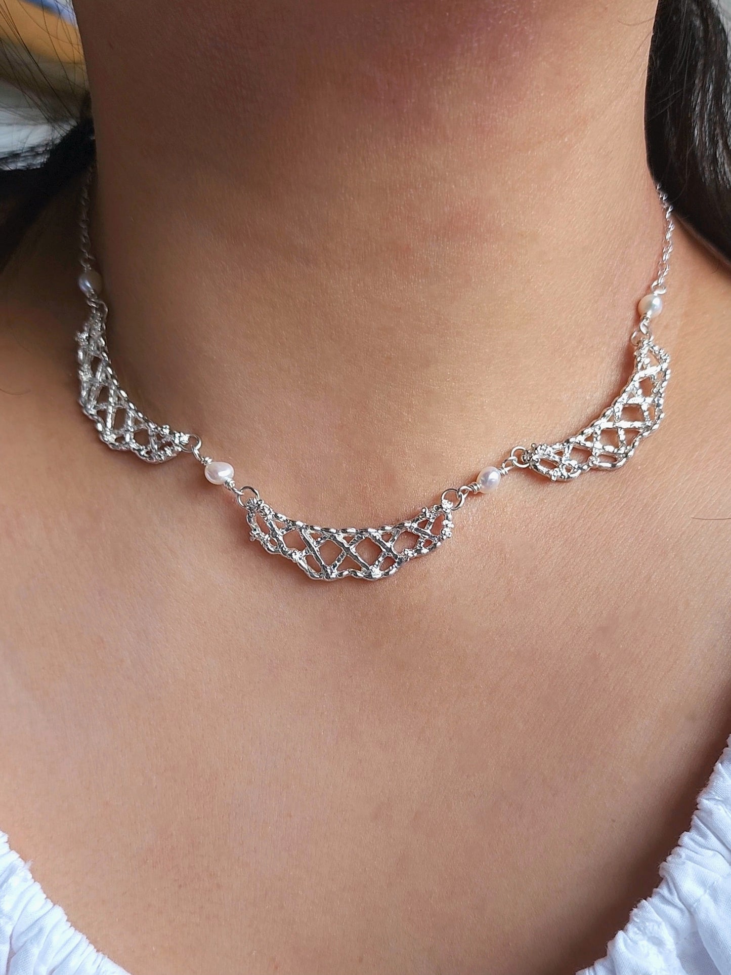 A close-up of a silver-toned necklace with a delicate openwork pattern and small pearl-like accents, worn on a person's neck.