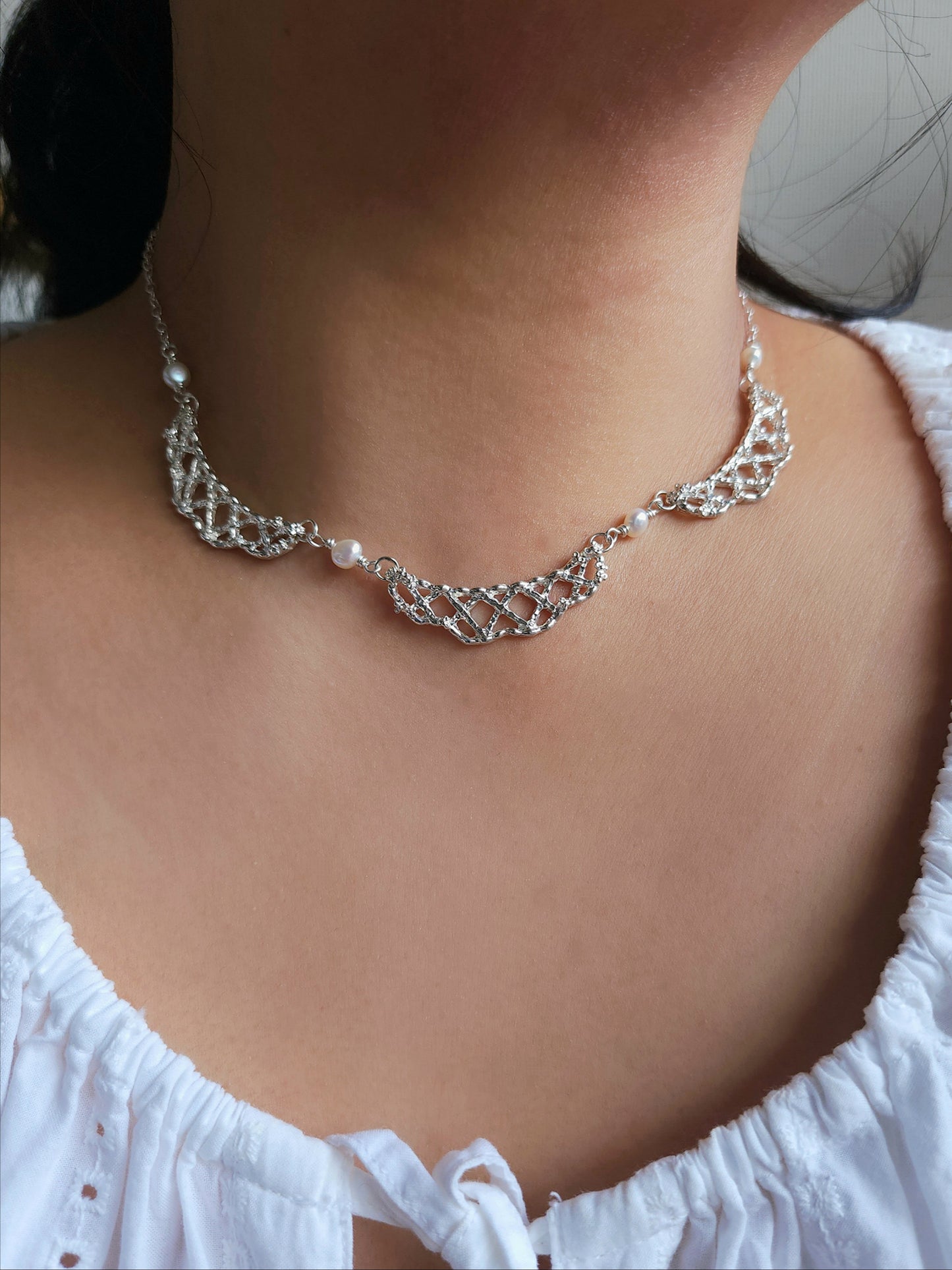 A close-up of a person's neck and upper chest wearing a delicate silver-toned choker necklace with a wavy openwork design and small pearl-like accents.