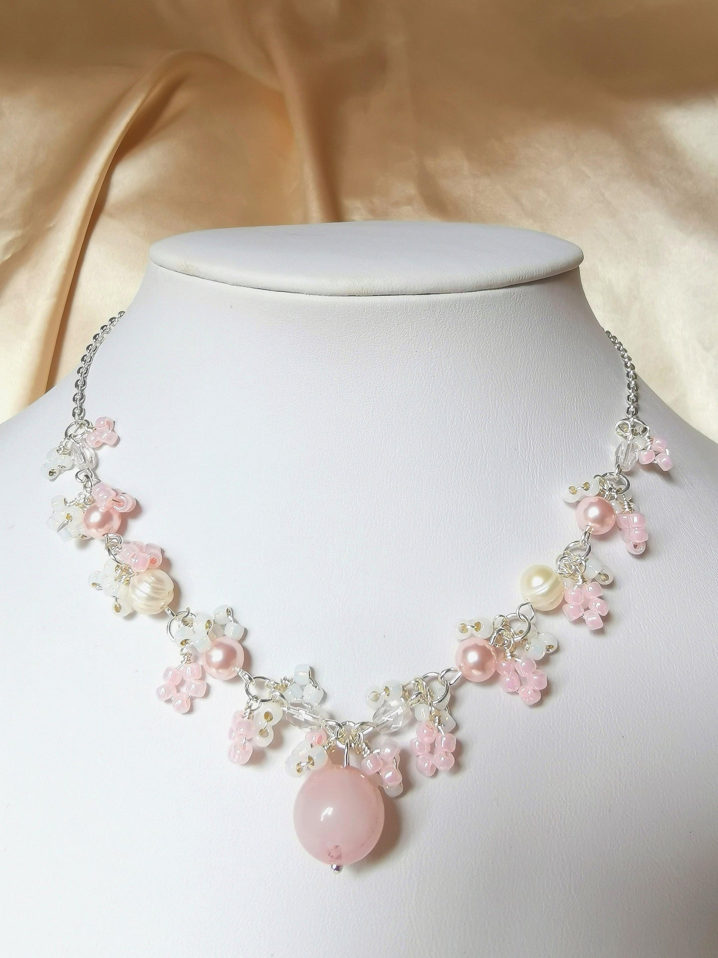 This is a handcrafted necklace where a rose quartz sphere pendant fans out to pink crystal pearls and white freshwater pearls, framed by white and pink seed bead details. The rest of the necklace is connected by silver-plated chain.